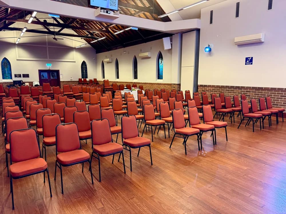 Empty church or auditorium with rows of orange chairs, brick wainscoting, arched stained glass windows, ceiling fans, air conditioning units, and a high wooden ceiling.