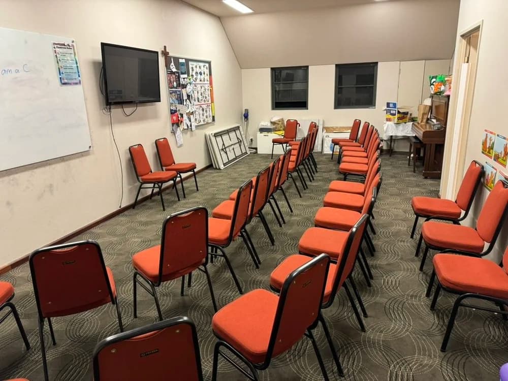 A small meeting room with orange chairs arranged in rows facing a whiteboard and a wall-mounted television. The room has two windows, a corner with storage boxes and a small table with a piano. There are colorful posters on the wall and some objects on the table and cabinets.