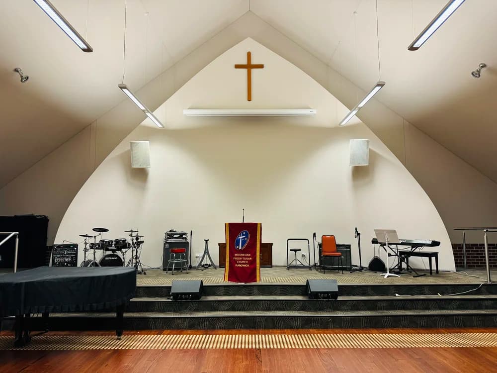 Stage inside a church with musical instruments including drums, keyboard, and microphones. A red banner with yellow trim displays the text 'Indonesian Presbyterian Church Randwick.' A cross is mounted on the wall behind the stage, and ceiling lights are visible.